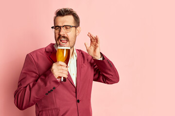 Portrait of stylish emotive man in a suit posing with glass of lager beer isolated on pink...