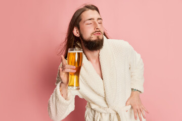 Portrait of emotive funny man in bathrobe posing with glass of lager beer isolated on pink...