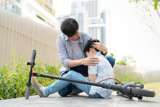Young Asia boy fell from electric scooter bike sitting on the floor and feeling pain and get injury on his knee in the public garden. Father holds the child's head in his arms and gives encourage.