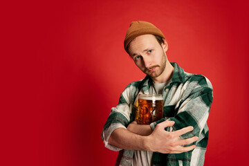 Portrait of young man in casual checkered shirt posing, hugging glass with beer isolated over red background. Favorite drink