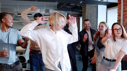 Young energetic people dancing in office of modern IT company having fun. Group of multiethnic employees having corporate party smiling with blonde happy woman in center. Corporate event concept.