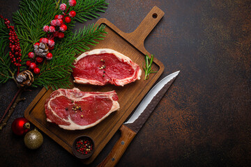Two raw uncooked meat beef steaks on wooden cutting board with knife and seasonings on dark rustic background with Christmas festive decoration from above with space for text