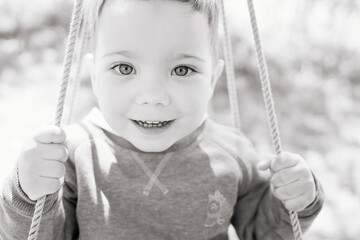 Happy three year old boy swings on a wooden swing on a sunny day in the countryside. Black and white. The concept of active childhood, games in the fresh air and harmonious education