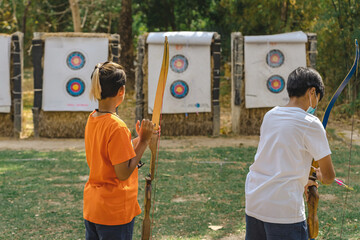 Back view of Asian boy wear face mask aims archery bow and arrow to colorful target in shooting...