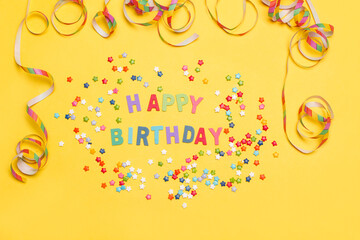 Cartel Feliz cumpleaños junto a estrellitas y confeti sobre un fondo amarillo liso. Vista superior y de cerca. Copy space