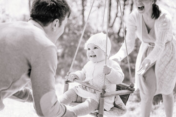 Stylish young parents swings their happy one-year-old daughter,  on a wooden swing on a sunny day in the countryside, black and white image, the concept of child care, safety for children.