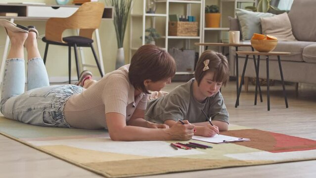 Teenage girl with ds and her loving mom lying on colorful rug together drawing