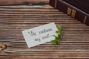 Biblical quote handwritten on lined paper with green plant and a closed holy bible book on wooden background. A close-up. Psalm 23, Christian verse, restoration and hope in the God Jesus Christ.