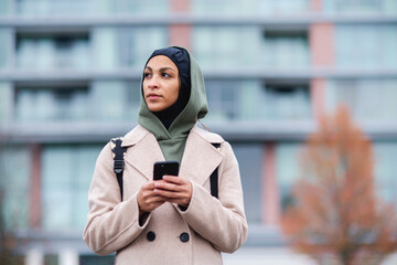 Portrait of young muslim woman in coat, standing outdoor in city, scrolling smartphone.