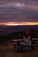 Naklejka na ściany i meble Young woman is sitting with thermos at bench and look at sunset mountains view