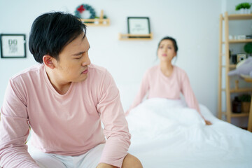 Obraz na płótnie Canvas Unhappy - stressful Asian man sitting on the bed while his wife shouting at him. Critical or serious arguing between husband and wife about a problem in family.