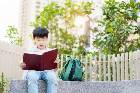 Cheerful Happy Asian Little Boy Sitting On The Bench And Reading A Book Or Fairy Tale At The Public Park Alone.