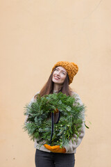 Smiling young woman in ginger hat and mittens holding Christmas wreath with pine and fir branches, mistletoe berries.