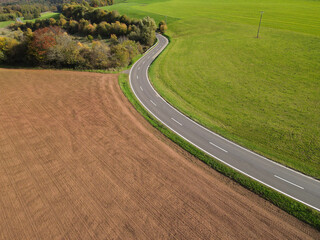 Aerial view of a long country road between plowed agricultural fields, trees and meadow on a warm day in autumn