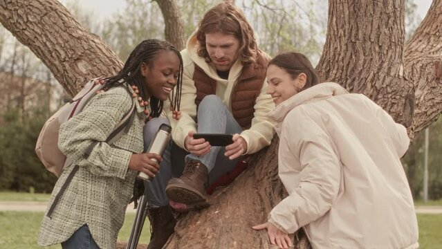 Young man sitting on tree trunk in park showing funny video on smartphone to his two female friends spending time outdoors together at springtime