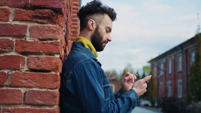 Portrait of focused young handsome man in jeans jacket texting on smartphone. Happy thoughtful guy chatting with girl while standing near wall. Buildings. Daytime. Autumn. Outside
