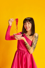 Portrait of young beautiful girl in bright pink dress eating pink noodles isolated on vivid yellow background. Concept of youth, beauty, fashion, lifestyle, emotions