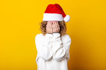 Curly young woman in a santa hat with her palms covering her face on a yellow background.
