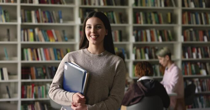 Portrait of attractive Hispanic student girl holding folder pose standing in campus library with schoolmates on background. College or university education, professional skill, knowledge, studentship