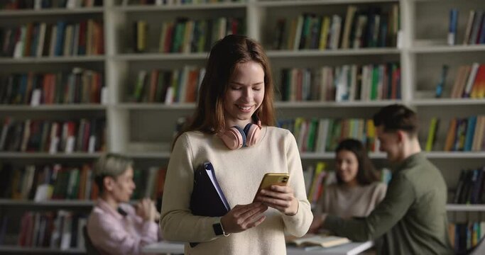 Young attractive redhead student girl standing in college library with smartphone, read message, check class school schedule using mobile application. Generation Z modern technology usage, lifestyle