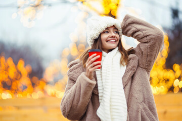 A pretty girl walks around the fair at Christmas, drinks a hot drink from a cup and looks away against the background of beautiful light decorations.