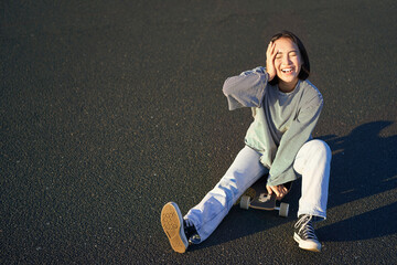 Happy beautiful korean teen girl sits on her skateboard, cruising on longboard, wearing casual...