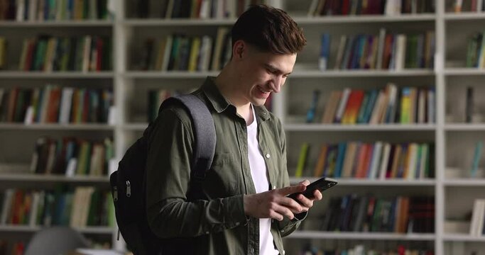 Highschool guy student with backpack holds modern smart phone check messages, chatting standing in university library. Generation Z using mobile application for studies, check class timetable online