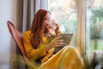Young woman with red hair reading on the tablet and drinking a hot coffee by the window at home