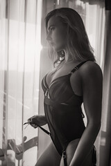 Young beautiful female pose in black body underwear lingerie by posing in living room  in sunset  light with big window on the background
