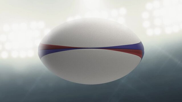 A seamlessly loop able animation of a white textured rugby ball with blue and red colour design element spinning and rotating on floodlit stadium background in the night time