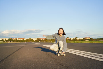 Carefree skater girl on her skateboard, riding longboard on an empty road, holding hands sideways...
