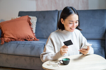 Smiling cute asian woman using credit card and smartphone, paying bills online, holding mobile...