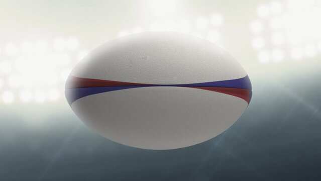 A seamlessly loop able animation of a white textured rugby ball with blue and red colour design element spinning and rotating on floodlit stadium background in the night time