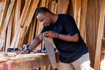 A young African carpenter saws wooden boards with a handsaw in a carpentry workshop.