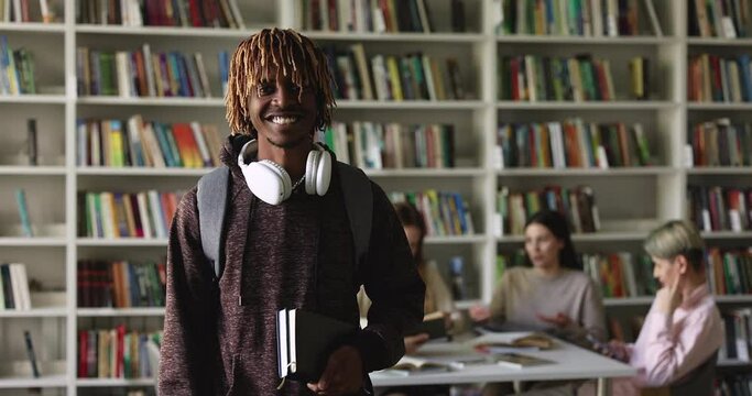 Smiling teenager African student guy with backpack and copybooks in hand posing in public or university library with mates on background. Excellent student portrait, high school education, knowledge