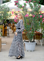 Attractive blonde Caucasian girl in a plaid midi dress, walking around in a flowery location. Pink flowering shrubs on a summer playground in a restaurant. Fashion and beauty concept