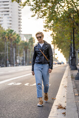 Adult 35s years old lesbian woman with sunglasses, leather jacket and blue jeans, walk along the street. Urban modern lifestyle in Barcelona city downtown in golden hour.