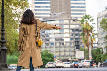 Fototapeta na wymiar Adult 35s years old businesswoman with long hair, sunglasses and trench coat catch a taxi in the downtown with modern buildings. Urban scene of Barcelona city.
