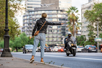 Adult 35s years old lesbian woman with sunglasses, leather jacket and blue jeans, catch a taxi on the street. Urban modern lifestyle in Barcelona city downtown in golden hour.