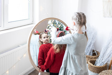 Cute sisters standing in front of mirror and preparing for holiday. Girls celebrating Christmas together at home