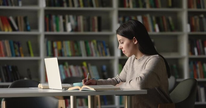 Attractive focused student girl preparing for session, college admission or university exams using laptop, writing information, studying seated at desk in library, side view. Education, modern tech