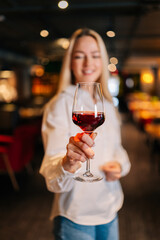 Vertical portrait of cheerful blonde woman holding glass of red wine standing in restaurant with luxury interior, looking away. Front view of cute female posing at cafe, blurred background.