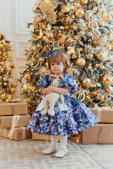 A little girl in a pretty blue dress is standing by the Christmas tree hugging a soft toy