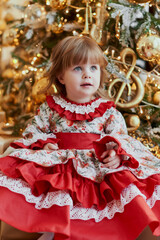 A little girl in a pretty red dress sits near the Christmas tree