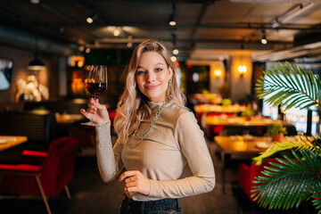 Portrait of smiling pretty woman holding glass of red wine standing in restaurant with luxury...