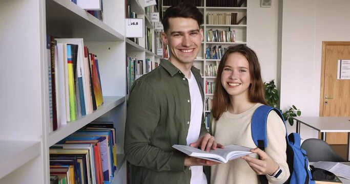 Attractive guy and girl, friendly schoolmates preparing for common task, standing in library posing with textbook in hands smile look at camera. Friendship, get higher education, successful studies