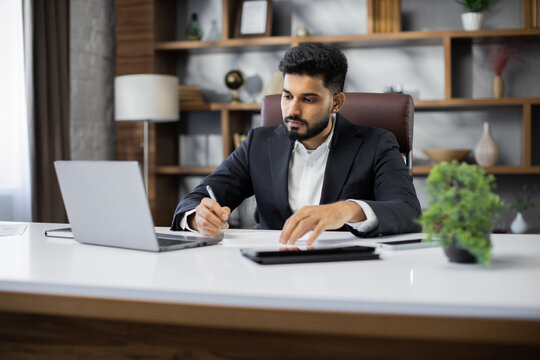 Confident businessman wearing suit writing notes or financial report, sitting at desk with laptop, focused serious man working with paper documents, student studying online, research work