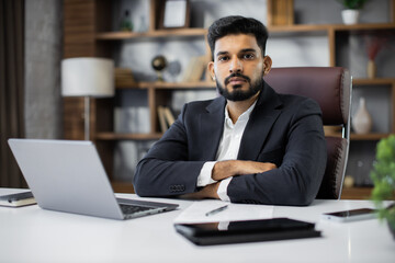 Obraz premium Confident young middle eastern businessman sitting at work table at modern office, typing on computer keyboard, sending emails to his business partners, working on marketing research, copy space