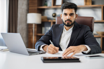 Fototapeta na wymiar Young bearded male accountant working from modern office using laptop while holding a document and pen in his hands and sign new deal or writing report.