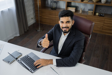 Obraz premium High angle view of stylish bearded businessman works on laptop, data analysis and creative designer, looking at computer showing thumb up. Digital entrepreneur working on e-commerce startup project